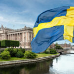 The building of the Swedish Parliament (Riksdag) and the Riksbank Bridge over the Lilla Vartan Strait with the national flag of Sweden in the foreground.