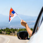 Woman holding Serbia flag from the open car window driving along the serpentine road in the mountains. Concept